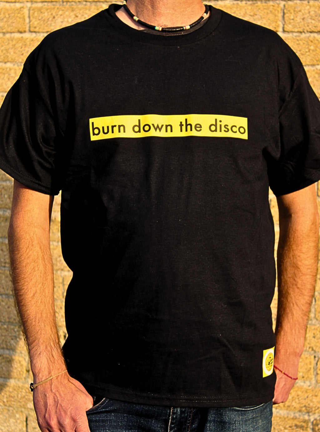 Person wearing a black t-shirt with yellow text 'burn down the disco' against a brick wall.