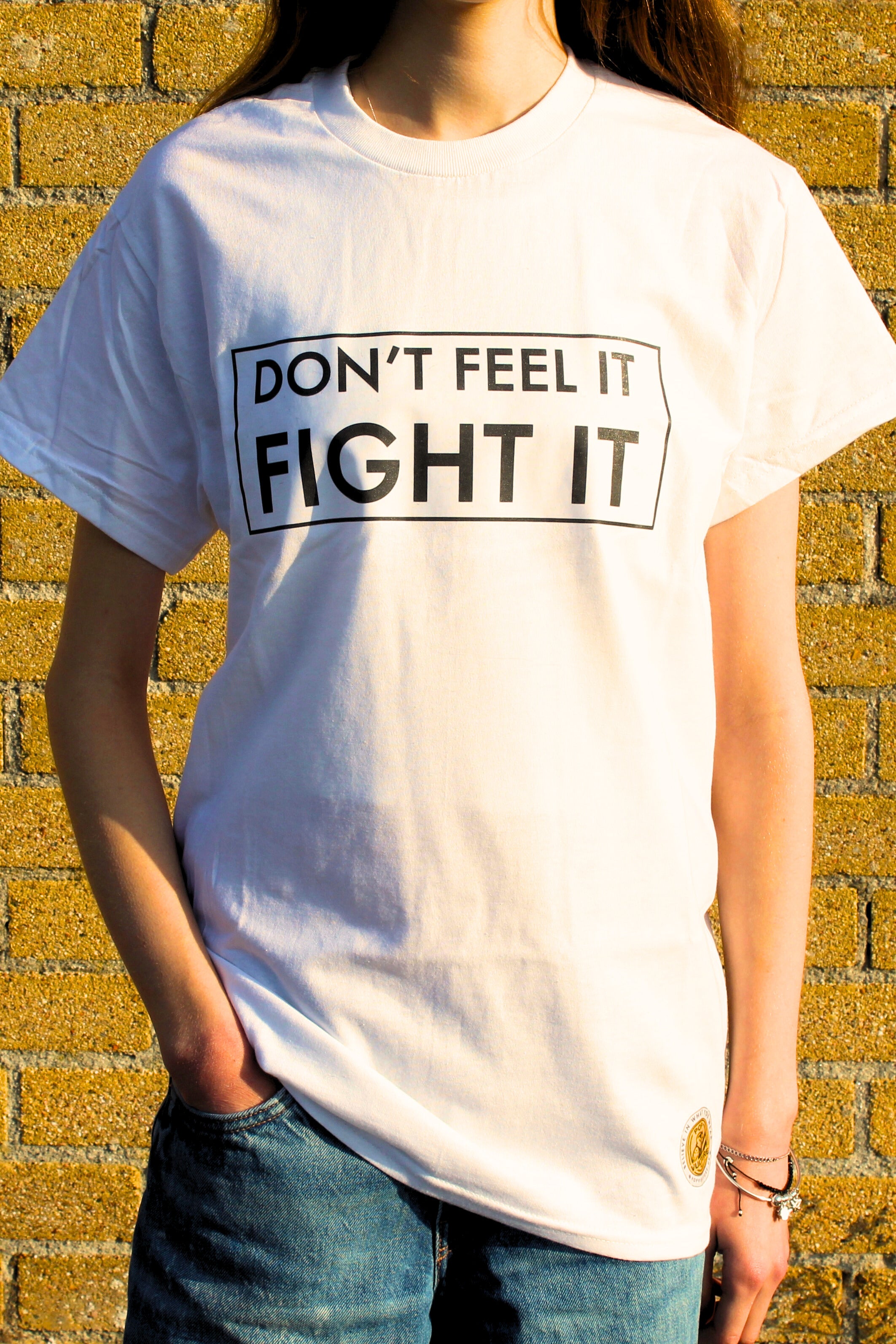 Person wearing a white t-shirt with 'DON'T FEEL IT FIGHT IT' text against a brick wall.