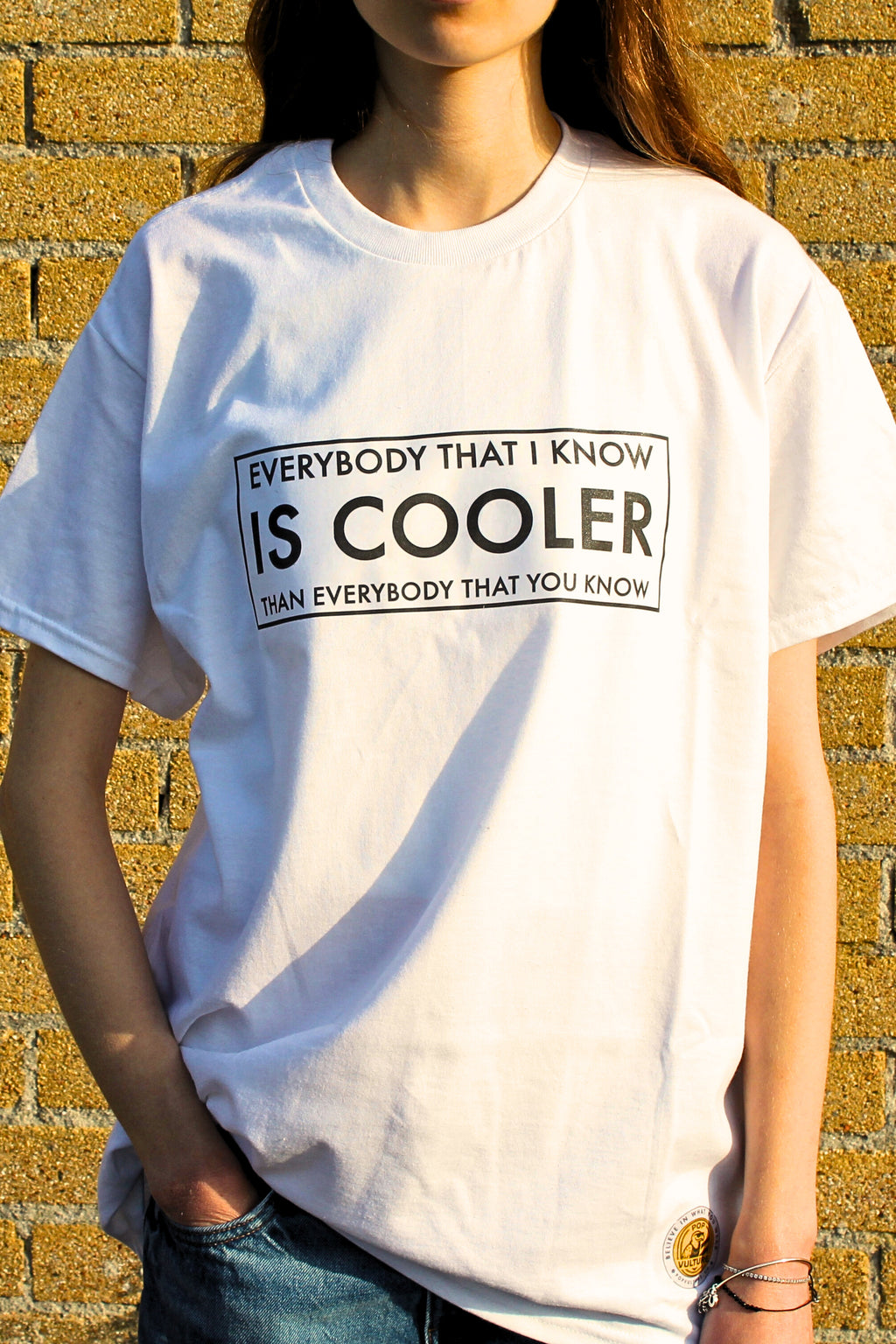 Person wearing a white t-shirt with a humorous text design against a brick wall.