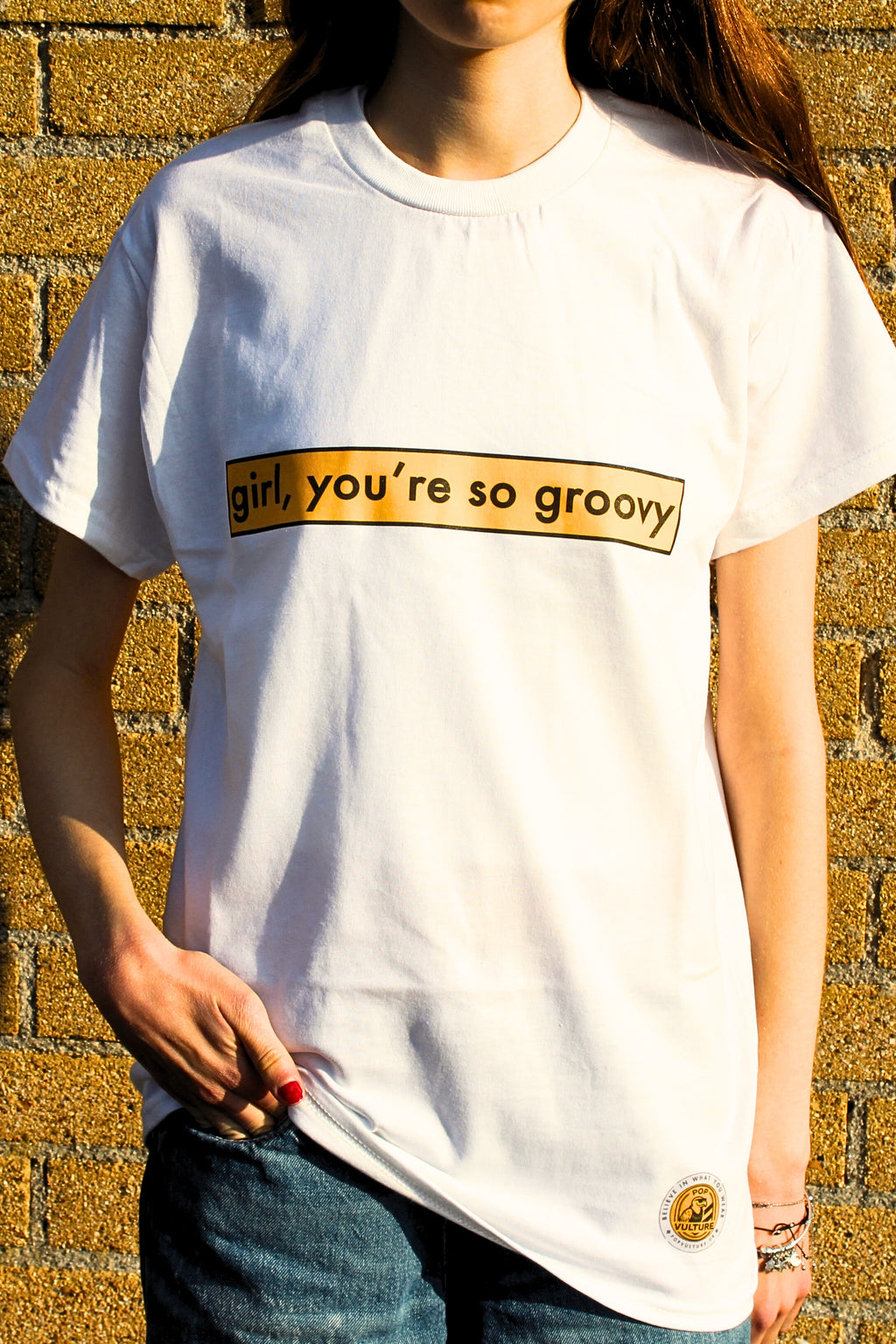 Person wearing a white t-shirt with 'girl, you're so groovy' text against a textured wall.