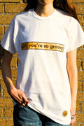 Person wearing a white t-shirt with 'girl, you're so groovy' text against a textured wall.