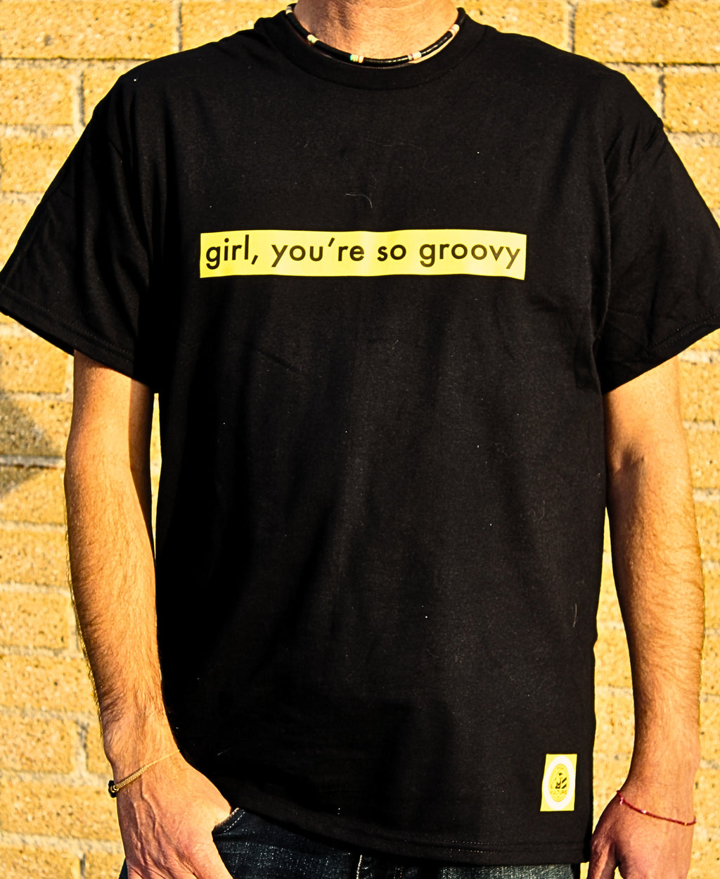 Person wearing a black t-shirt with yellow text 'girl, you're so groovy' against a brick wall.
