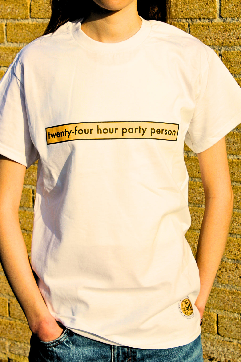 Person wearing a white t-shirt with 'twenty-four hour party person' text against a brick wall.