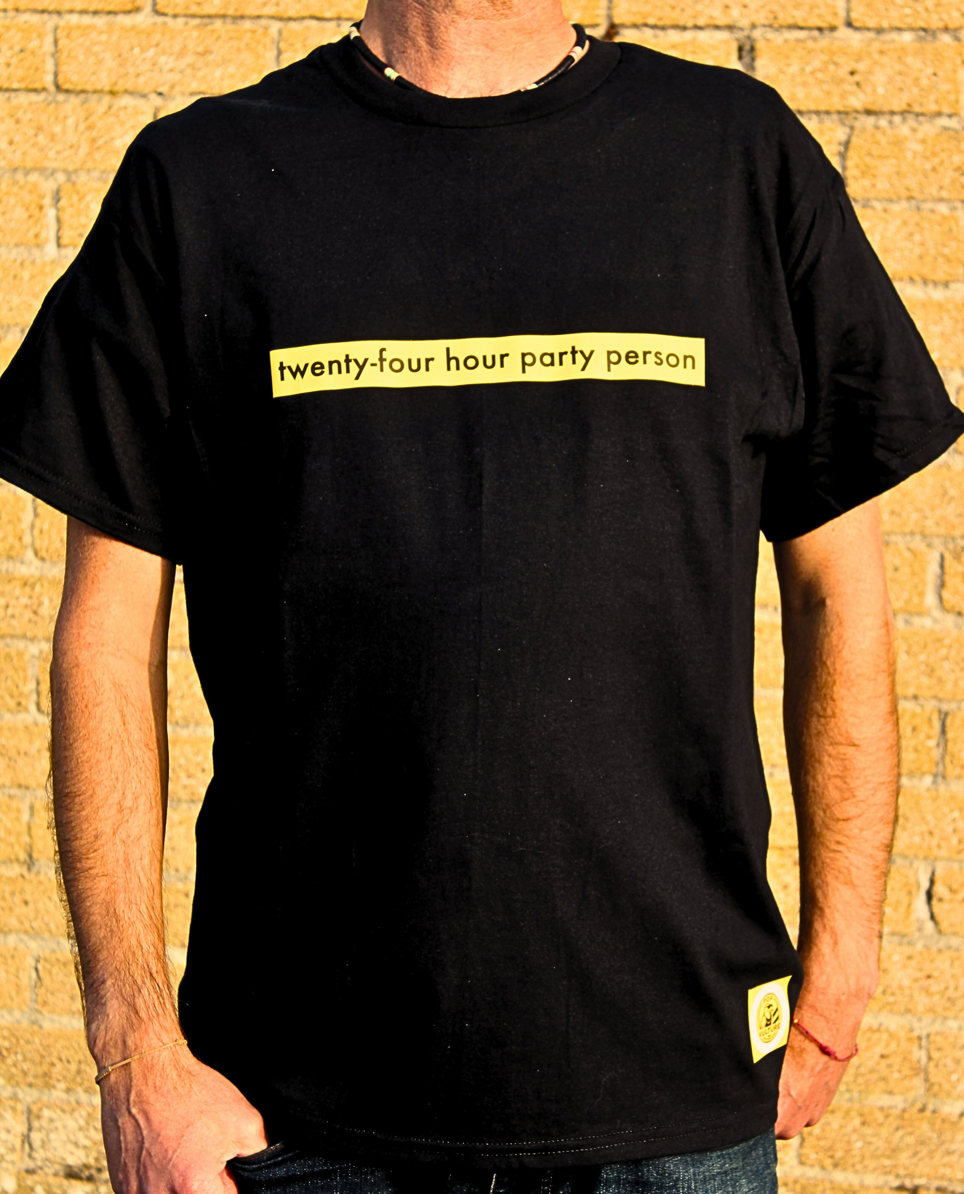 Person wearing a black t-shirt with yellow text 'twenty-four hour party person' against a brick wall.